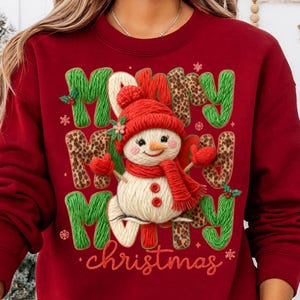 May include: A red sweatshirt with a festive Christmas design. The design features the words "Merry Christmas" with a snowman wearing a red hat, scarf, and mittens. The letters are green and leopard print.