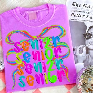 May include: A lilac-colored t-shirt with the word "Senior" in a colorful, layered design. The word is in a rainbow of colors, with a bow design above it. A newspaper clipping is in the background.