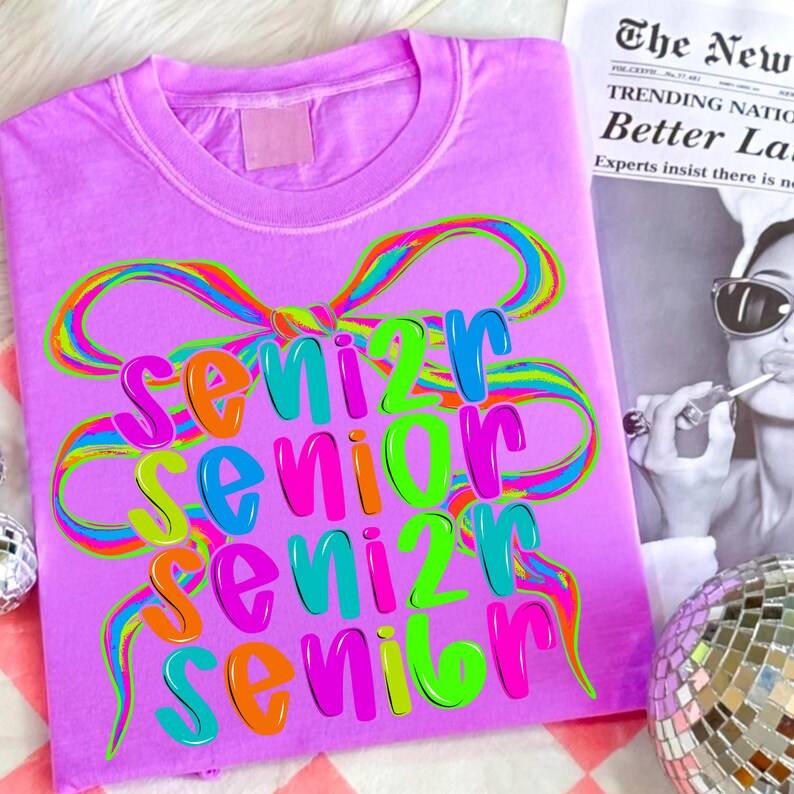May include: A lilac-colored t-shirt with the word "Senior" in a colorful, layered design. The word is in a rainbow of colors, with a bow design above it. A newspaper clipping is in the background.