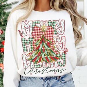 May include: White sweatshirt with a festive Christmas design. The design features the words "MERRY CHRISTMAS" in a mix of green and red plaid and floral patterns, with a Christmas tree and bow.