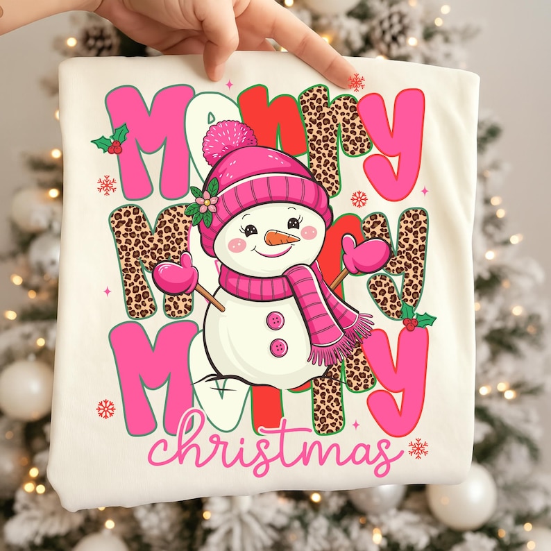 May include: Cream-colored t-shirt with a festive Christmas design. The design features the words "Merry Christmas" in pink and leopard print, along with a snowman wearing a pink hat, scarf, and mittens. Snowflakes and holly accents complete the holiday theme.
