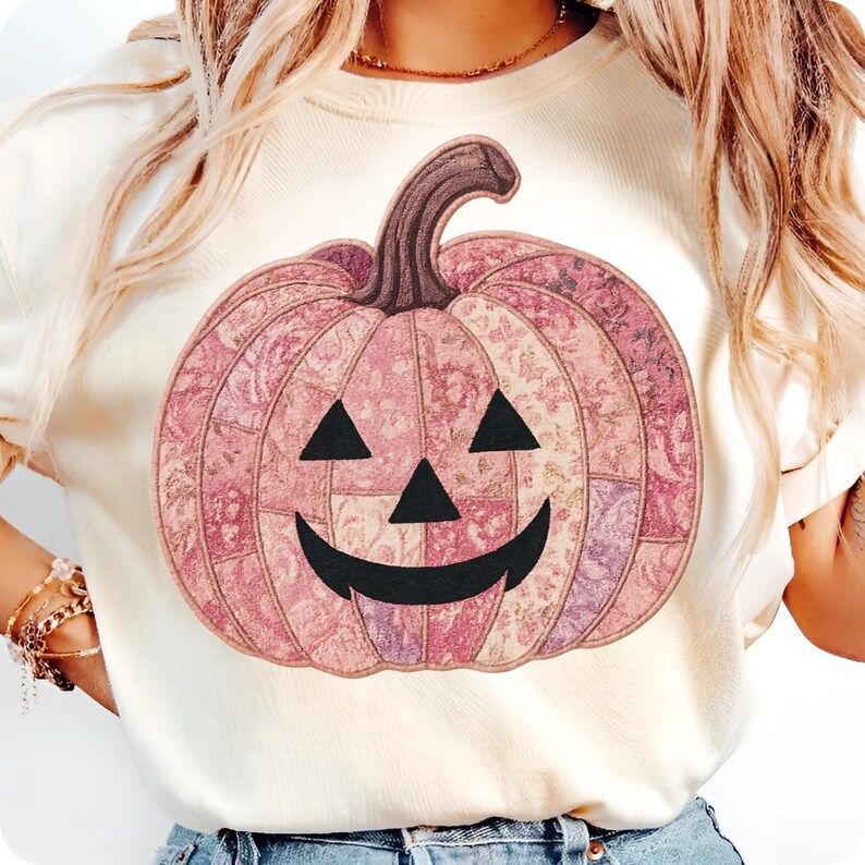 K&ouml;nnte beinhalten: Cremefarbenes T-Shirt mit einem rosa K&uuml;rbisdesign. Der K&uuml;rbis hat ein Patchworkmuster in Rosa- und Lilat&ouml;nen, mit einem schwarzen K&uuml;rbisgesicht und einem braunen Stiel. Ein festliches Halloween-Grafik-T-Shirt.