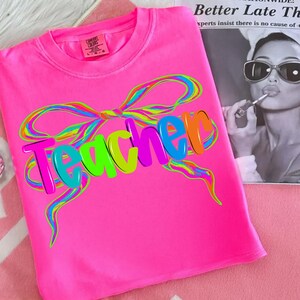 Op de afbeelding: Felroze t-shirt met het woord "Teacher" in kleurrijke 3D-letters en een regenboogstrikontwerp. Het shirt heeft een ronde hals en is gemaakt van zacht materiaal. Een merklabel is zichtbaar.
