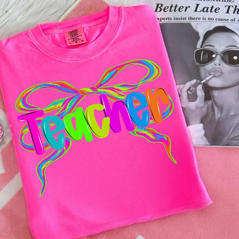 Op de afbeelding: Felroze t-shirt met het woord "Teacher" in kleurrijke 3D-letters en een regenboogstrikontwerp. Het shirt heeft een ronde hals en is gemaakt van zacht materiaal. Een merklabel is zichtbaar.