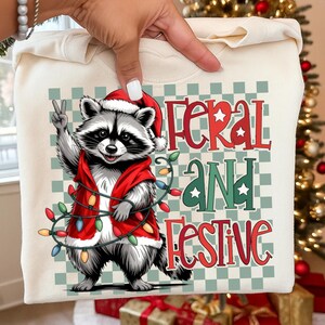May include: Cream-colored sweatshirt featuring a cartoon raccoon wearing a Santa hat and a red vest, holding a string of Christmas lights. The text "FERAL AND FESTIVE" is displayed in red and green. The raccoon is giving a peace sign.