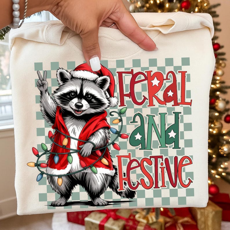 May include: Cream-colored sweatshirt featuring a cartoon raccoon wearing a Santa hat and a red vest, holding a string of Christmas lights. The text "FERAL AND FESTIVE" is displayed in red and green. The raccoon is giving a peace sign.