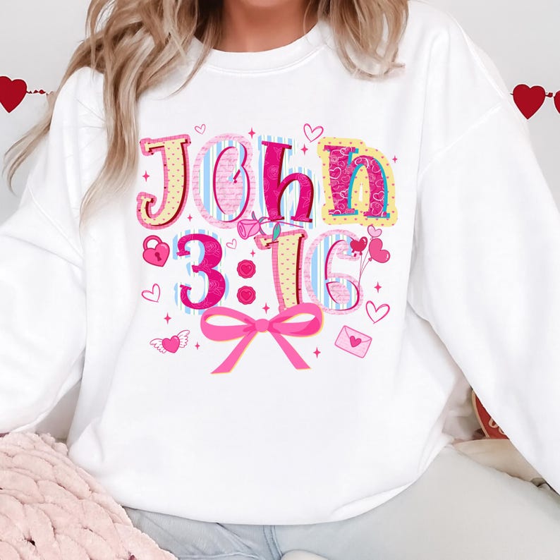 May include: White sweatshirt featuring the text "John 3:16" in a colourful, playful design. The graphic includes hearts, a bow, and other Valentine's Day themed illustrations in pink, blue, and yellow, creating a festive look.