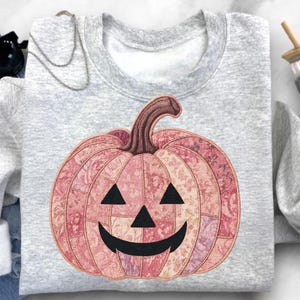 K&ouml;nnte beinhalten: Ein hellgraues Sweatshirt mit einem rosa floralen K&uuml;rbisdesign. Der K&uuml;rbis hat einen braunen Stiel und schwarze Dreiecksaugen sowie einen l&auml;chelnden Mund. Das Design ist auf der Vorderseite des Sweatshirts zentriert, ideal f&uuml;r Halloween.