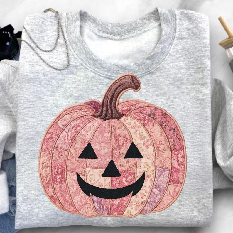 K&ouml;nnte beinhalten: Ein hellgraues Sweatshirt mit einem rosa floralen K&uuml;rbisdesign. Der K&uuml;rbis hat einen braunen Stiel und schwarze Dreiecksaugen sowie einen l&auml;chelnden Mund. Das Design ist auf der Vorderseite des Sweatshirts zentriert, ideal f&uuml;r Halloween.