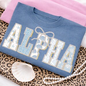 May include: A blue sweatshirt featuring the word "ALPHA" in white, adorned with seashells, starfish, and pearls. The sweatshirt is folded on pink fabric and a leopard print cloth, with a seashell and pearl necklace.