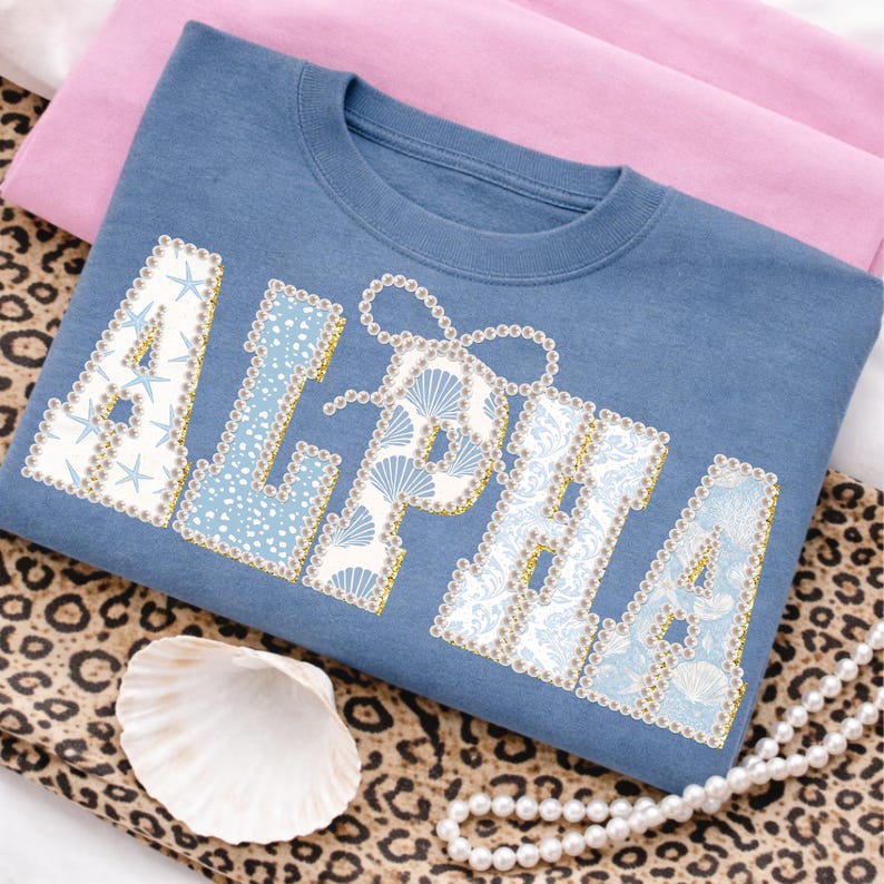 May include: A blue sweatshirt featuring the word "ALPHA" in white, adorned with seashells, starfish, and pearls. The sweatshirt is folded on pink fabric and a leopard print cloth, with a seashell and pearl necklace.