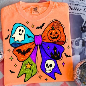 May include: Orange t-shirt with a large, glittery Halloween bow design. The bow features a ghost, pumpkin, bats, stars, a skull, and a haunted house. The shirt has a Comfort Colors tag.