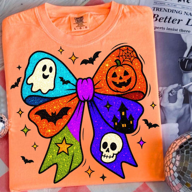 May include: Orange t-shirt with a large, glittery Halloween bow design. The bow features a ghost, pumpkin, bats, stars, a skull, and a haunted house. The shirt has a Comfort Colors tag.