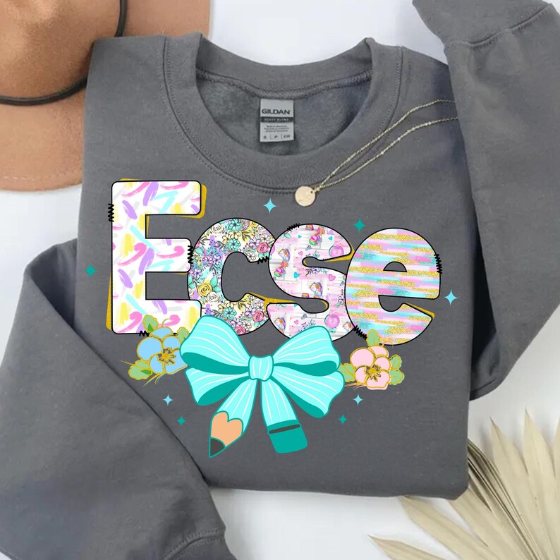 May include: A dark gray sweatshirt with the word "Ecse" in colorful, patterned letters. The design includes a turquoise bow, a pencil, and floral accents. A gold necklace is visible.