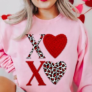 Może przedstawiać: Jasnor&oacute;żowa bluza z literami "X" i czerwonym motywem serca. Jedna "X" ma wz&oacute;r w cętki, druga jest w jednolitym kolorze czerwonym. Czerwone serce jest dziane, a drugie serce ma wz&oacute;r w cętki.