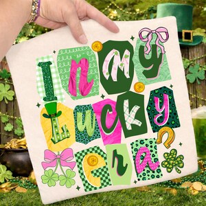 May include: A cream-colored pillow with the words "I'm in my lucky era" in a colorful, whimsical design. The design includes green, pink, and gold accents, with shamrocks, bows, and a leprechaun hat.