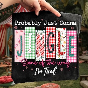 Op de afbeelding: Zwart t-shirt met de tekst "Probably Just Gonna JINGLE Some of the way I'm Tired." Het woord "JINGLE" staat in kleurrijke, feestelijke blokken met zuurstokken, ruitjes en kerstboomontwerpen.