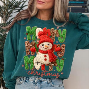 May include: Teal green sweatshirt with a snowman graphic and the words "Merry Christmas." The snowman wears a red hat, scarf, and mittens. The letters "M" and "Y" are green, and the letters "R" and "S" have a leopard print.