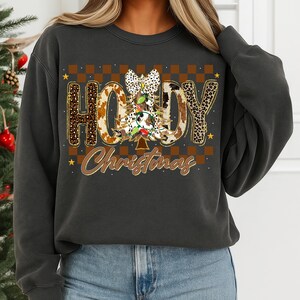 May include: Dark gray sweatshirt with a festive design. The design features the word "Howdy" in a mix of leopard and cow print, with a Christmas tree and the word "Christmas" in a brown script. The background has a brown and tan checkered pattern.