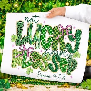 May include: A white print with the words "not LUCKY simply BLESSED" in green, with pink bows and shamrock accents. The text "Roman 4:7-8" is at the bottom. The print is held by a person.