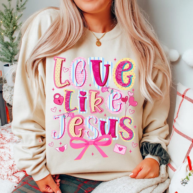 May include: Beige sweatshirt with the words "Love Like Jesus" in colorful, decorative lettering. The design includes hearts, a lock, a bow, and an envelope. The sweatshirt is a casual, comfortable style.