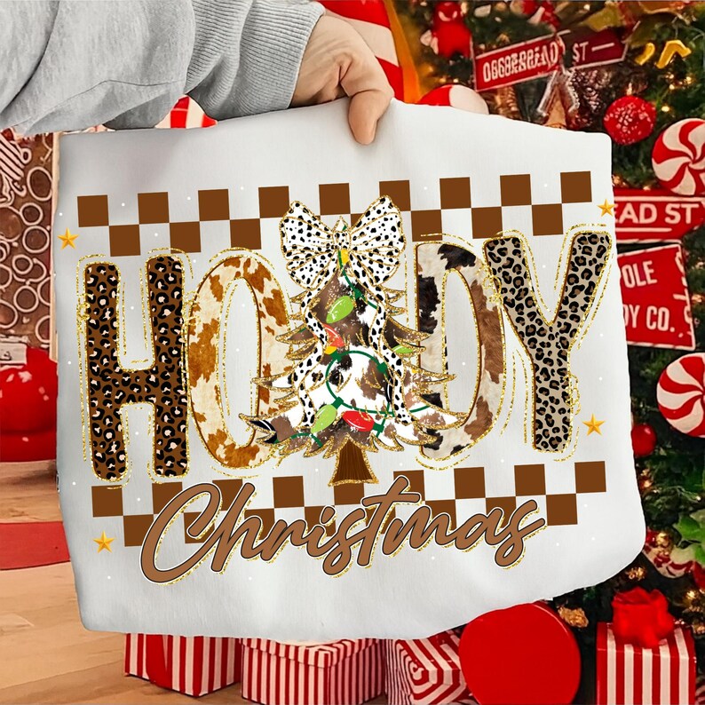 May include: White sweatshirt with the words "HOLLY Christmas" in a brown, cowhide, and leopard print design. The letter "O" is a Christmas tree with a cowhide pattern, a bow, and Christmas lights. The sweatshirt is held up by a person.