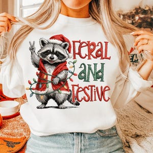 May include: White sweatshirt featuring a raccoon wearing a Santa hat and a red vest, holding up a peace sign. The text "Feral and Festive" is printed in red and green. A string of colorful Christmas lights is wrapped around the raccoon.