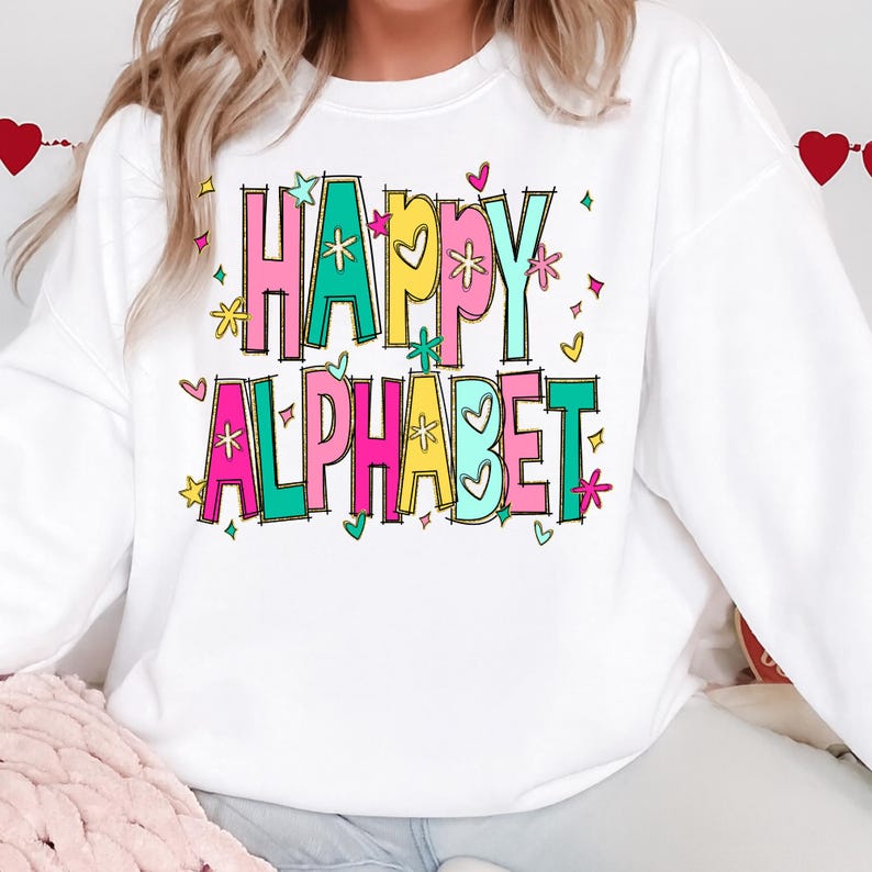K&ouml;nnte beinhalten: Wei&szlig;es Sweatshirt mit den Worten "HAPPY ALPHABET" in bunten Blockbuchstaben. Die Buchstaben sind schwarz umrandet und mit Rosa, Gelb und T&uuml;rkis gef&uuml;llt. Das Design umfasst Herzen, Sterne und florale Akzente.