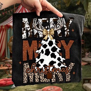 May include: Black t-shirt with a Christmas design. The design includes the words "MERRY CHRISTMAS" in a mix of fonts and patterns, including cow print, leopard print, and a Christmas tree shape. A gold bow and antlers are also part of the design.