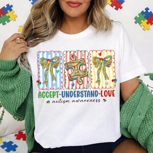 May include: White t-shirt with a colourful graphic promoting autism awareness. The design features three panels with bows and a puzzle piece, along with the words "ACCEPT-UNDERSTAND-LOVE" and "autism awareness."
