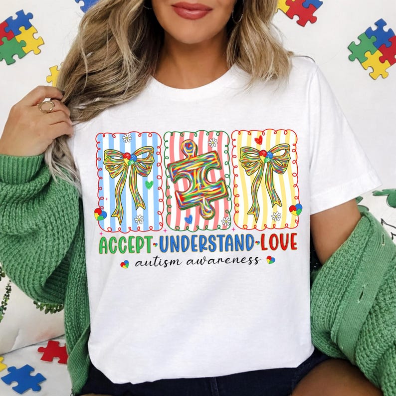 May include: White t-shirt with a colourful graphic promoting autism awareness. The design features three panels with bows and a puzzle piece, along with the words "ACCEPT-UNDERSTAND-LOVE" and "autism awareness."