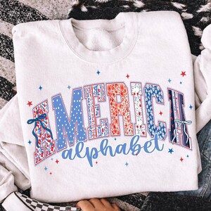 May include: White sweatshirt with the word "AMERICA" in a patriotic design. The letters are filled with floral and polka dot patterns in red, white, and blue. The word "alphabet" is written below in blue.