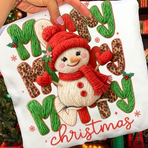 May include: White Christmas pillow featuring a snowman wearing a red hat, scarf, and mittens. The words "Merry Christmas" are embroidered in green, red, and leopard print. Snowflakes and holly accents complete the festive design.