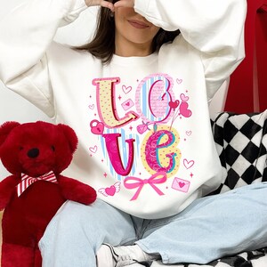 以下が含まれることがあります： カラフルな装飾文字で「LOVE」と書かれた白いスウェットシャツ。文字はピンク、黄色、青で、ハートとリボンのアクセントが付いています。蝶ネクタイをした赤いテディベアが左側にあります。