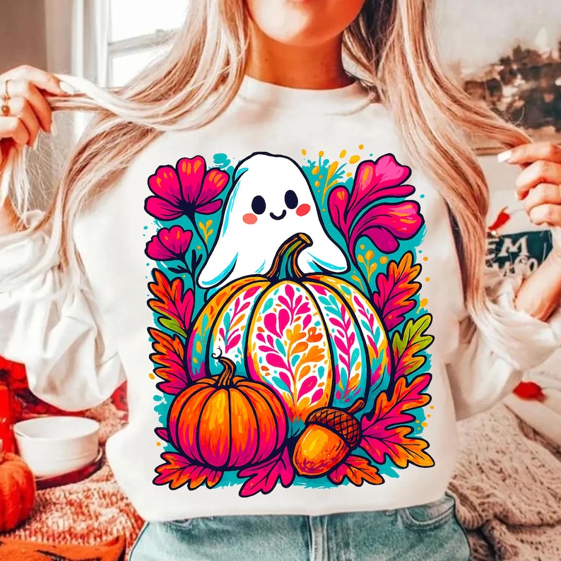 May include: White sweatshirt featuring a colorful Halloween design. The graphic includes a friendly ghost, a decorated pumpkin, a smaller pumpkin, and an acorn, surrounded by vibrant flowers and leaves. The design is in bright shades of orange, pink, and teal.