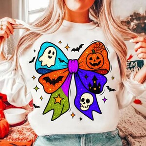 May include: White sweatshirt featuring a colorful Halloween bow design. The bow includes a ghost, pumpkin, bat, skull, and star. The bow is decorated with glitter and surrounded by bats and stars.
