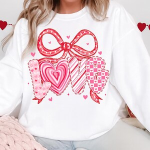 May include: White sweatshirt with a large red bow and the word "LOVE" in a heart-themed design. The letters are in pink and red with heart and stripe patterns. Small hearts are scattered around the design.
