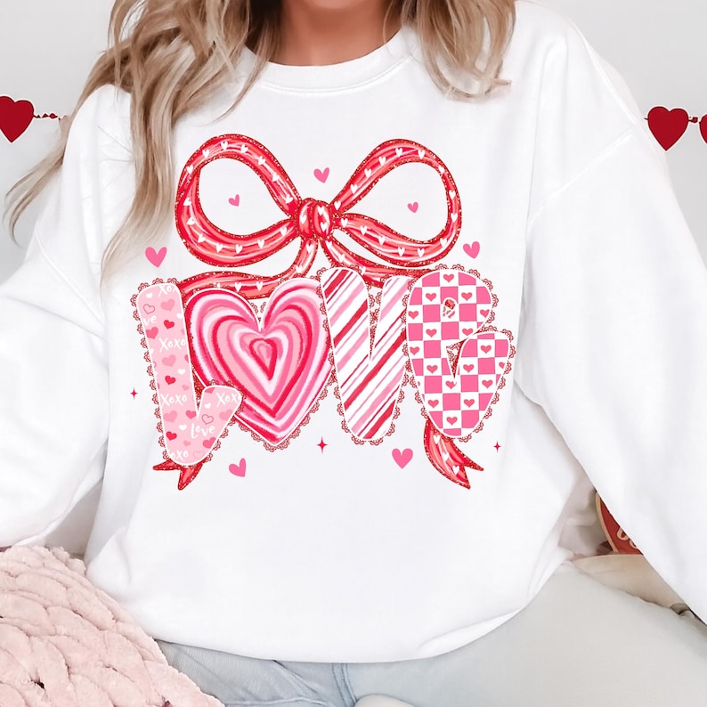 May include: White sweatshirt with a large red bow and the word "LOVE" in a heart-themed design. The letters are in pink and red with heart and stripe patterns. Small hearts are scattered around the design.