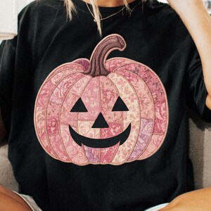 K&ouml;nnte beinhalten: Schwarzes T-Shirt mit einem rosa K&uuml;rbis-Design. Der K&uuml;rbis hat ein Patchwork-Muster in Rosa- und Lilat&ouml;nen mit einem geschnitzten Gesicht. Der Stiel ist braun. Eine Halloween-Grafik.