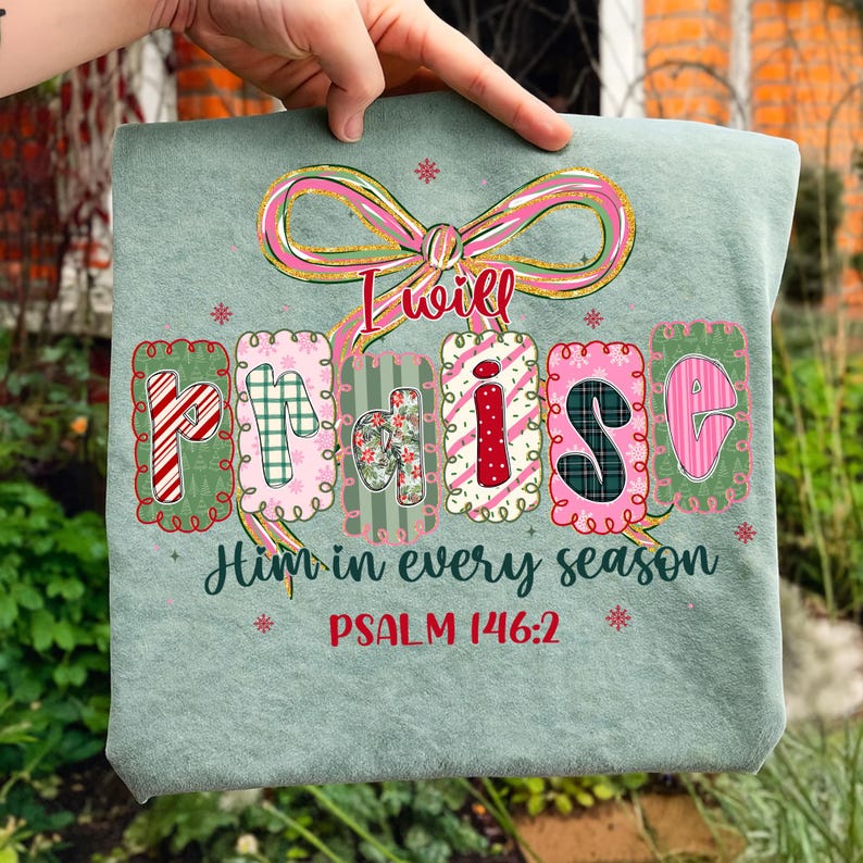 May include: A light green t-shirt with the words "I will praise Him in every season" and "PSALM 146:2" printed on it. The word "praise" is in colorful, decorative letters. A pink and gold bow is at the top.