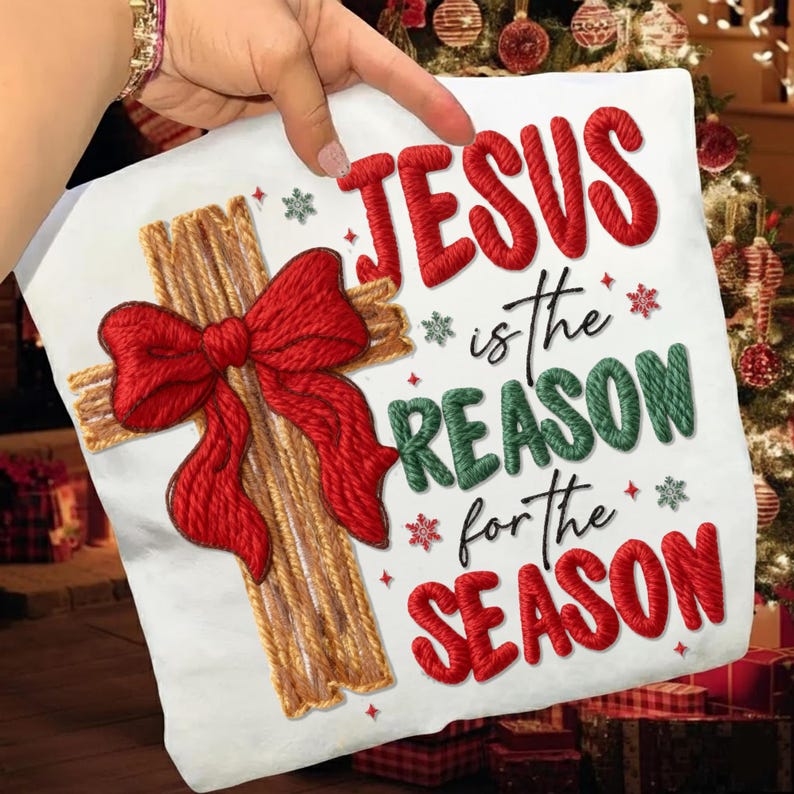 May include: White pillow with a cross design, a red bow, and the text "JESUS is the REASON for the SEASON" in red and green. The cross is made of brown yarn, and the bow is red.