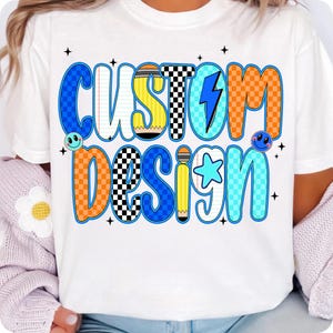 May include: White t-shirt featuring the words "CUSTOM DESIGN" in a colorful, retro design. The text incorporates blue, orange, yellow, and black checkered patterns, with a lightning bolt and star graphic. Smiley faces and stars accent the design.