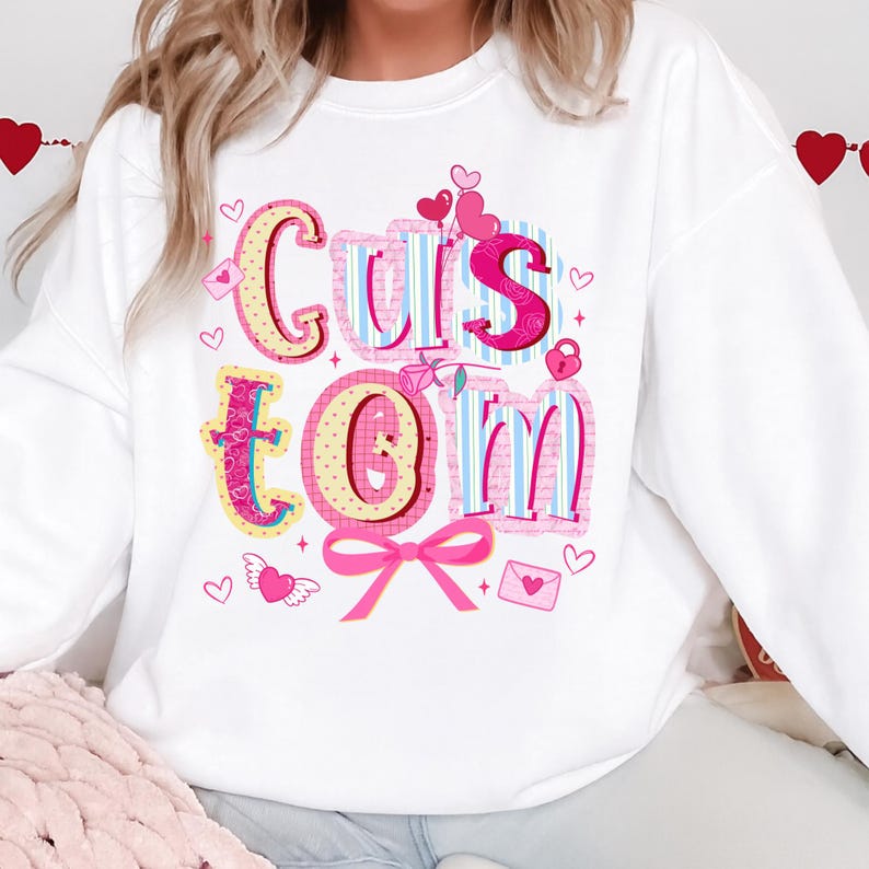May include: White sweatshirt with the word "Custom" in a colorful, Valentine's Day-themed design. The letters are decorated with hearts, bows, and other romantic elements. The sweatshirt is a casual, comfortable style.