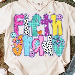 May include: Cream-colored t-shirt with the words "FIFTH GRADE" in a colorful, patchwork design. The text features various patterns, including leopard print, polka dots, stars, and a lightning bolt. Smiley faces and stars are also incorporated.