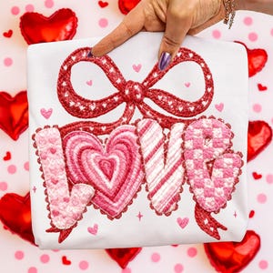 May include: White fabric with a red bow and the word "LOVE" embroidered in pink. The bow is decorated with white hearts. The letters are in various pink patterns, including hearts and stripes. Small pink hearts are scattered around the design.