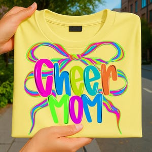 May include: A light yellow t-shirt with the words "Cheer Mom" in bright, colorful letters. The text is surrounded by a rainbow-colored ribbon design. The shirt is held up by a person.