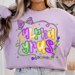 May include: A light purple sweatshirt with the words "Alpha Gras" in a colorful design. The text is outlined in yellow, green, and purple, with a bow and bead accents. The design includes gold glitter accents.