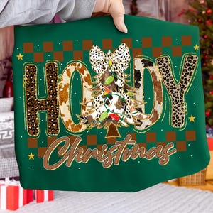 May include: Green Christmas-themed sweatshirt with the words "HO HOY Christmas" in a mix of leopard and cow print. A Christmas tree with lights and a bow is in the center. Gold accents and stars complete the design.