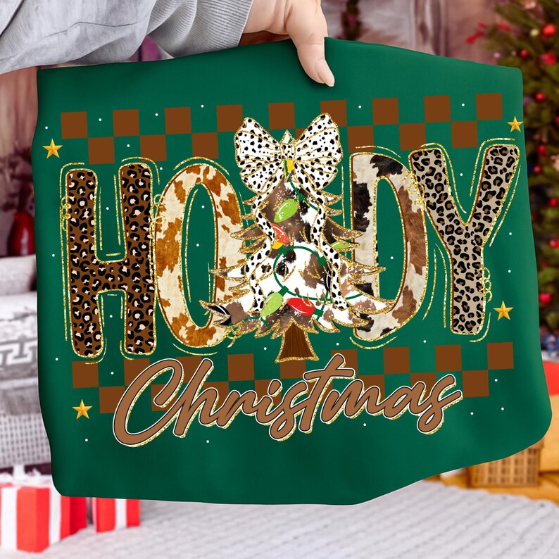 May include: Green Christmas-themed sweatshirt with the words "HO HOY Christmas" in a mix of leopard and cow print. A Christmas tree with lights and a bow is in the center. Gold accents and stars complete the design.