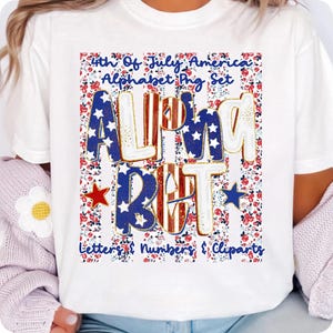May include: White t-shirt with a 4th of July design. The design features the word "ALPHABET" in a bold font with red, white, and blue stars and stripes. The shirt also includes the text "Letters & Numbers & Cliparts" against a floral background.